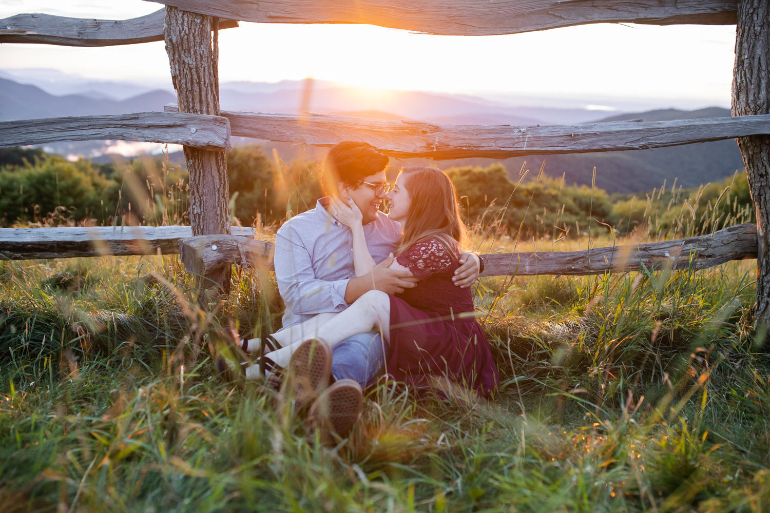 Boy and girl sitting in a field kissing again a wooden fence in front of a sunset at Max Patch NC.