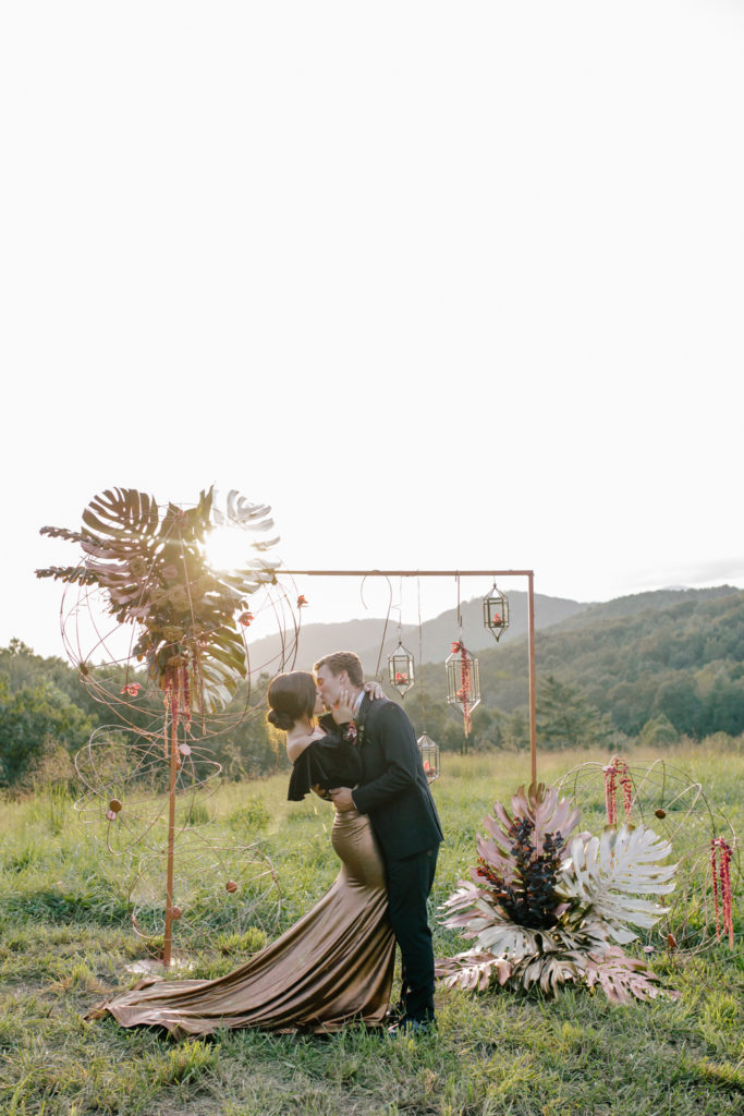 Bride, wearing a black shirt and gold skirt, kissing under a gold arbor at Mill Creek Farm in Hiddenite NC.