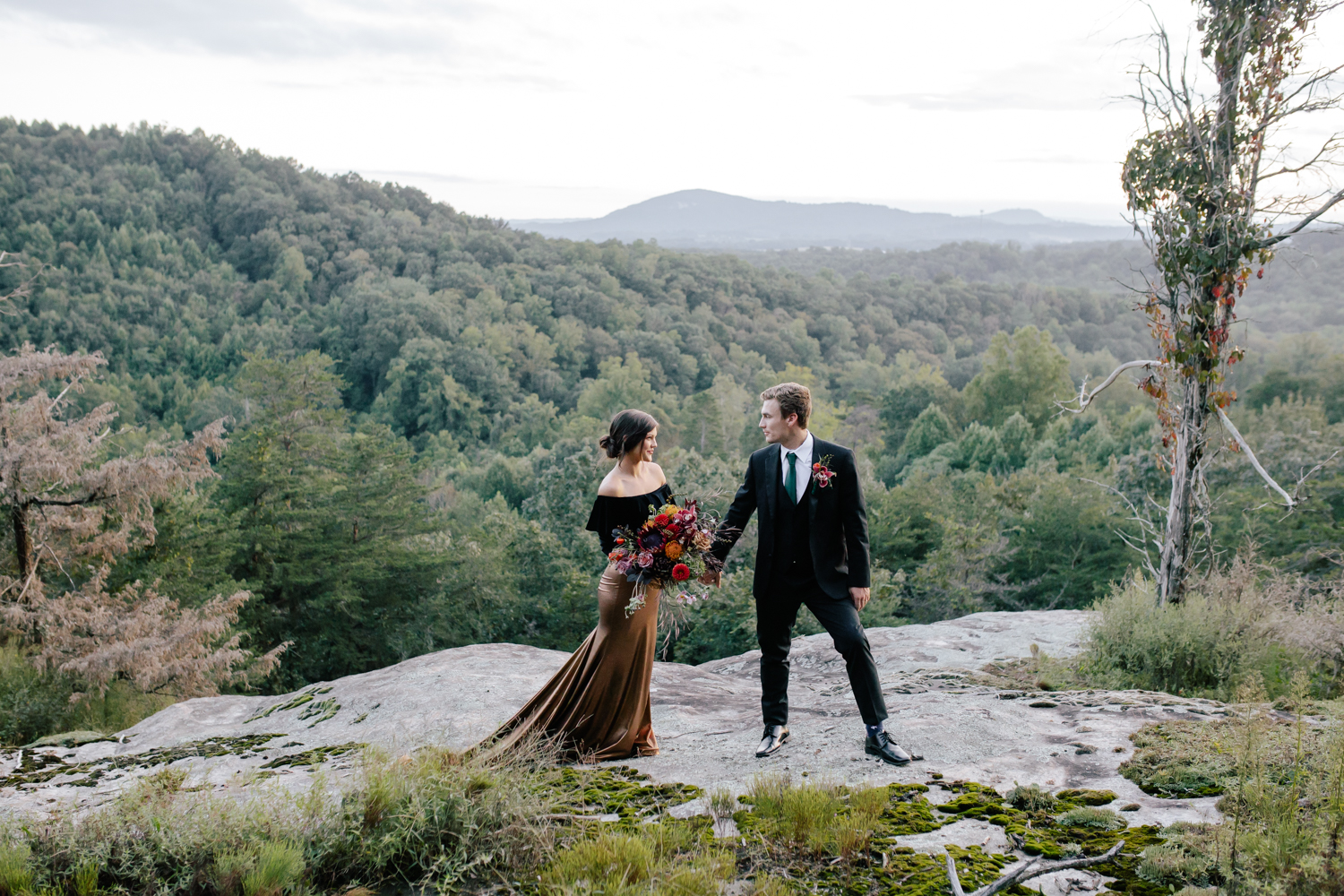 Bride, wearing a black shirt and gold skirt, holding hands with groom, wearing a black suit and green tie, walking on a mountain top at Mill Creek Farm in Hiddenite NC.