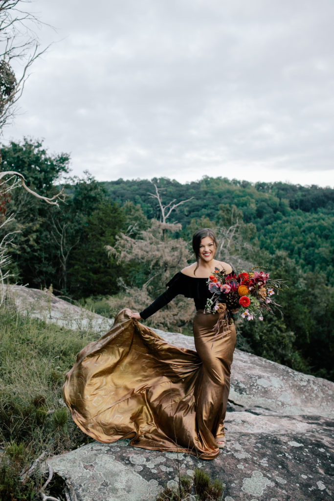 Bride dressed in a gold skirt and black shirt holding a red and purple bouquet of flowers on a mountain top at Mill Creek Farm in North Carolina.