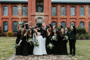 Wedding party, dressed in black dresses and suits, celebrate bride and groom at Providence Cotton Mill Wedding Venue near Charlotte NC.