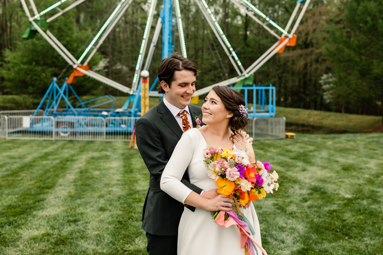 Groom, in a green suit, hugging his bride, in a white dress, in front of a blue ferris wheel.