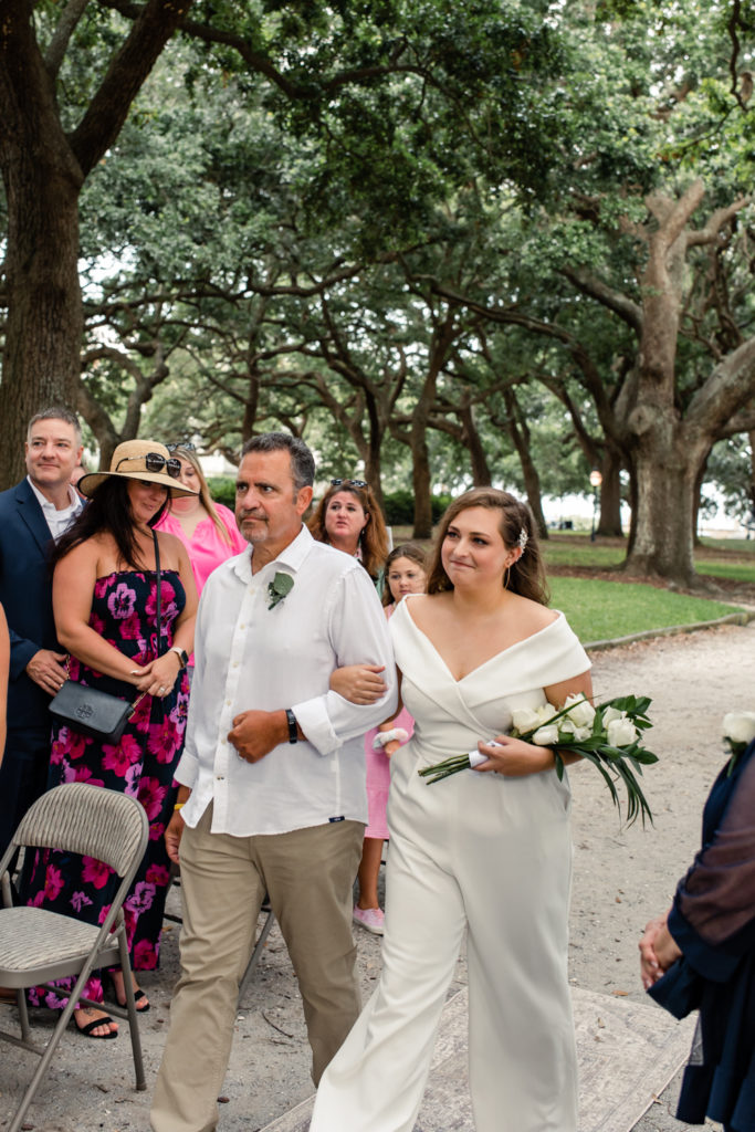 Bride, in white jumpsuit, walking with her dad, in white button down, down the aisle at the White Point Gardens in Charleston.