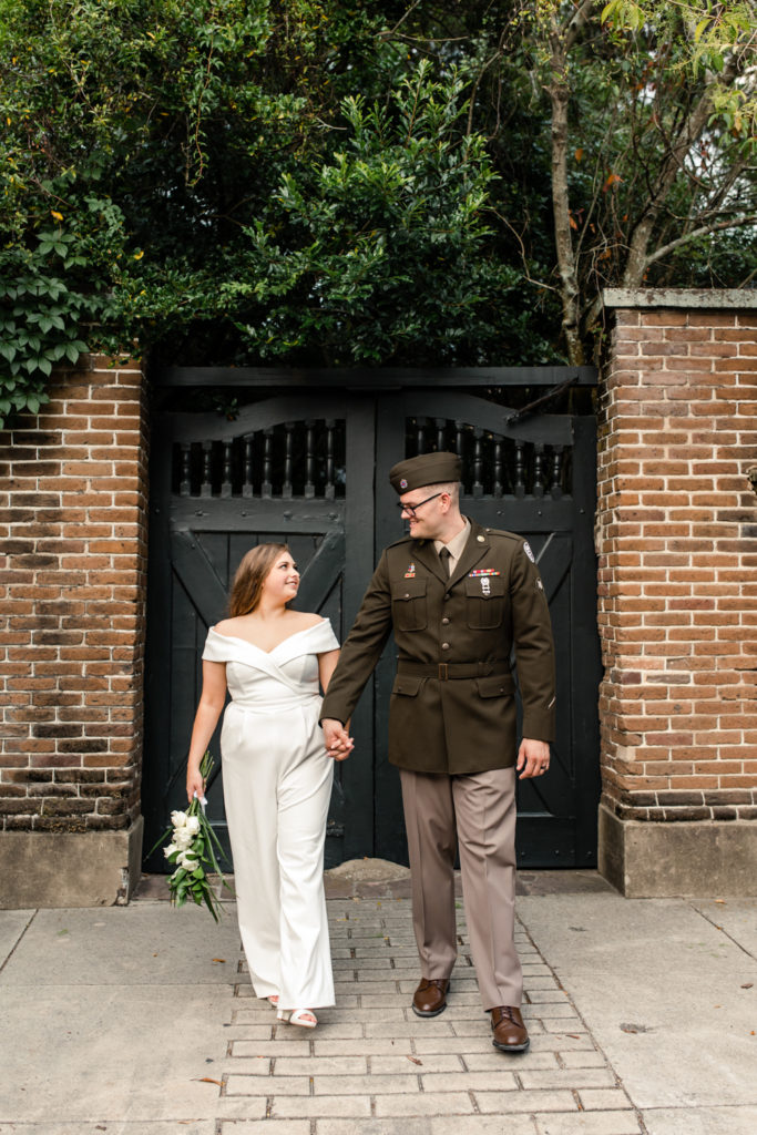 Bride, in white jumpsuit, walking with her groom, in green military uniform, at the White Point Gardens in Charleston.
