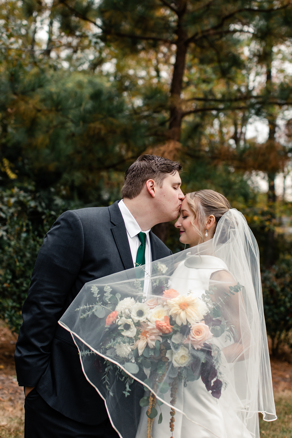 Groom, in white suit and green tie, kissing the forehead of his bride, in white dress and veil with green and red flowers at Carmel Country Club wedding venue. Photographed by Charlotte wedding photography.