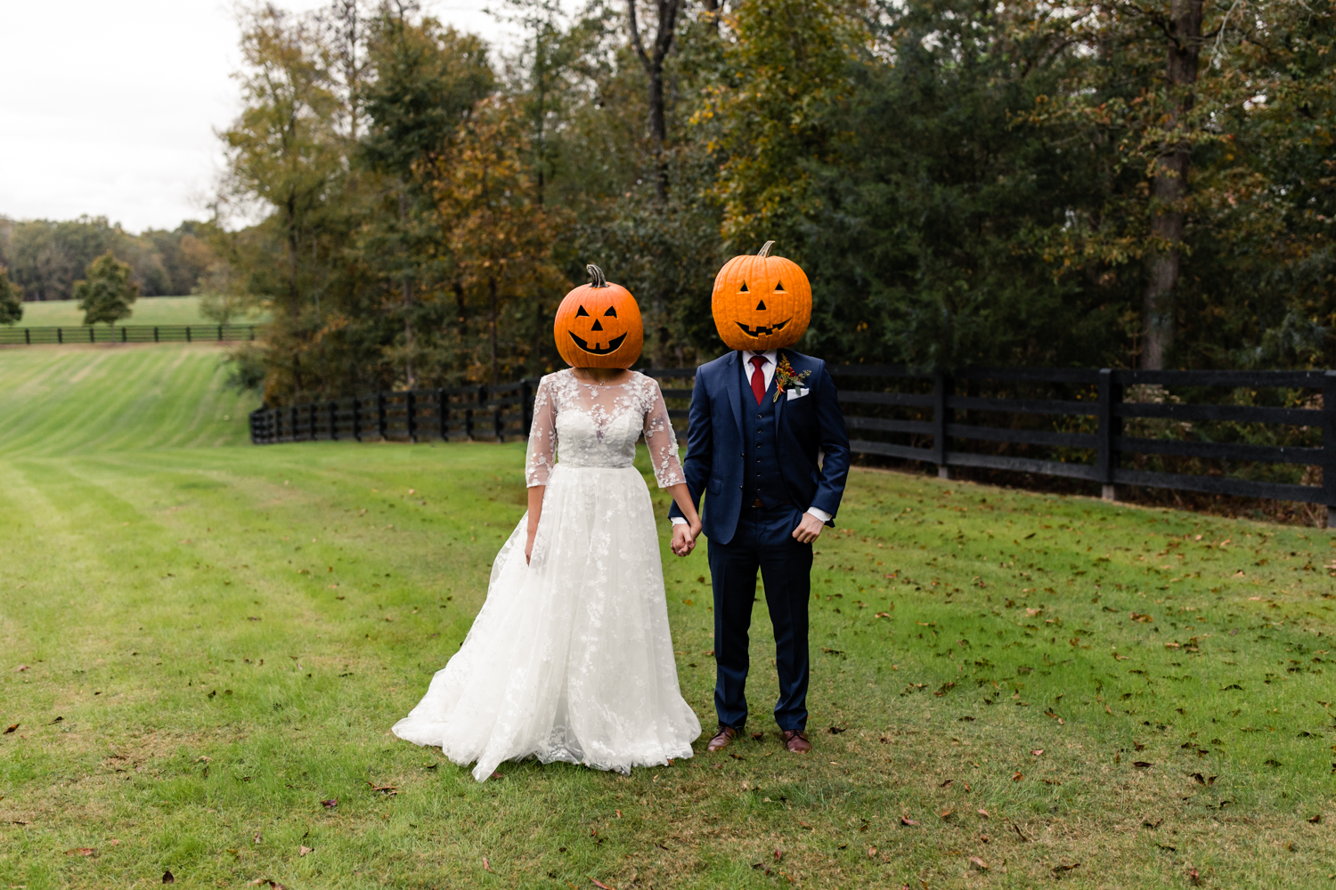 Bride, in white lace dress, holding hands with husband, in blue suit, wearing pumpkin heads at Rolling Hill Farms. Photographed by Charlotte wedding photography.