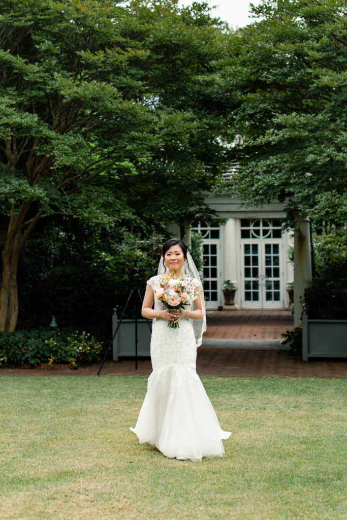 Bride in white lace gown walking down a green grass aisle holding pink and white bouquet at Daniel Stowe Botanical Garden wedding venue. Photographed by Charlotte wedding photography.