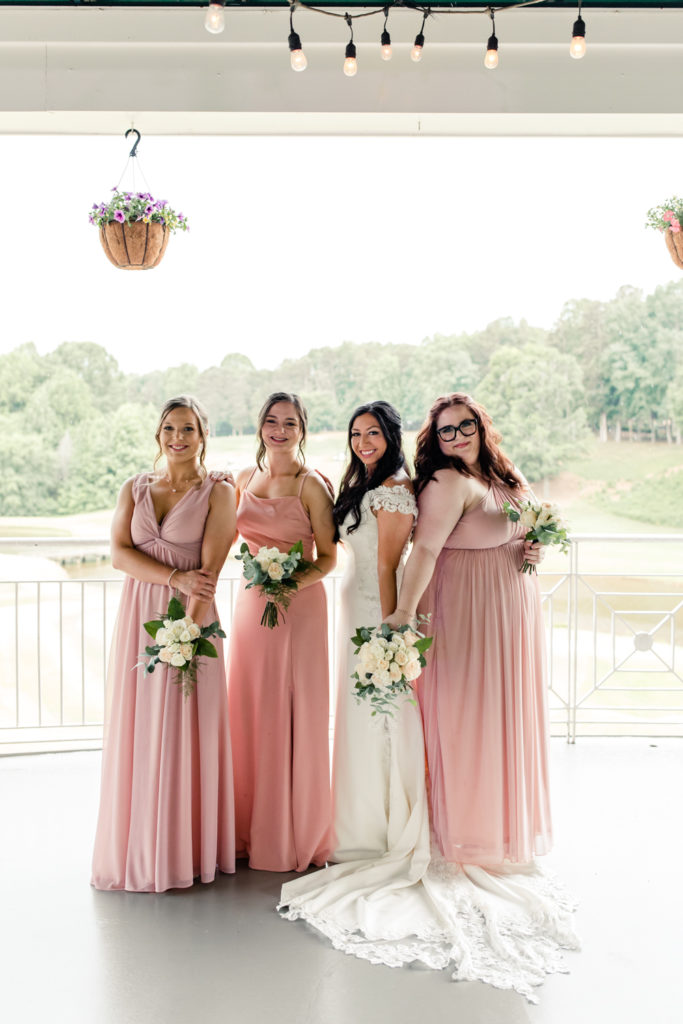 Bride in white lace dress standing with three bridesmaids in pink dresses holding white bouquets at Northstone Country Club in Charlotte. Photographed by Charlotte wedding photographer.
