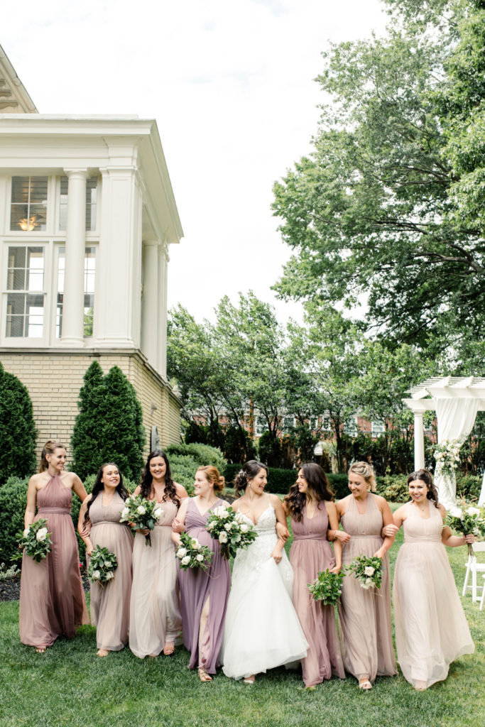 Bride in white dress walks with seven bridesmaids in pink dresses holding green and white bouquets at SePark Mansion Gastonia Wedding Venue. Photographed by Charlotte wedding photographer.