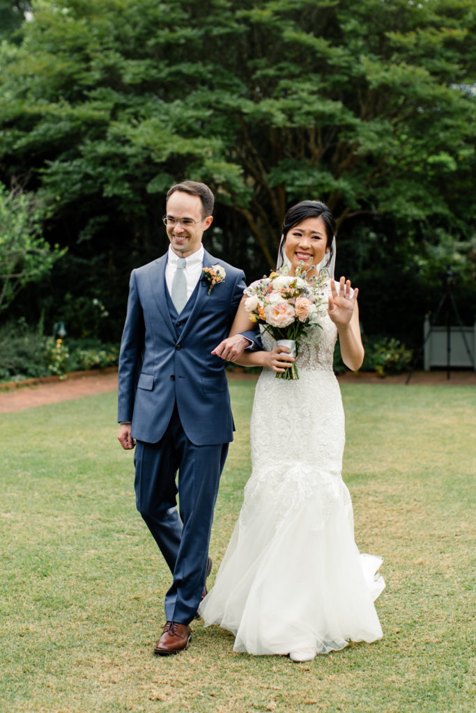 Bride in white lace dress and pink and white bouquet walking down aisle holding hands with groom in blue suit at Daniel Stowe Botanical wedding venue. Photographed by Charlotte wedding photography.