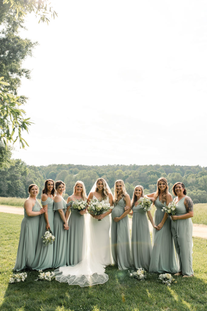 Bride in white wedding gown standing with eight bridesmaids, holding whit bouquets, in green field at The Meadows Raleigh wedding venue in Raleigh NC. Photographed by Charlotte wedding photographer.