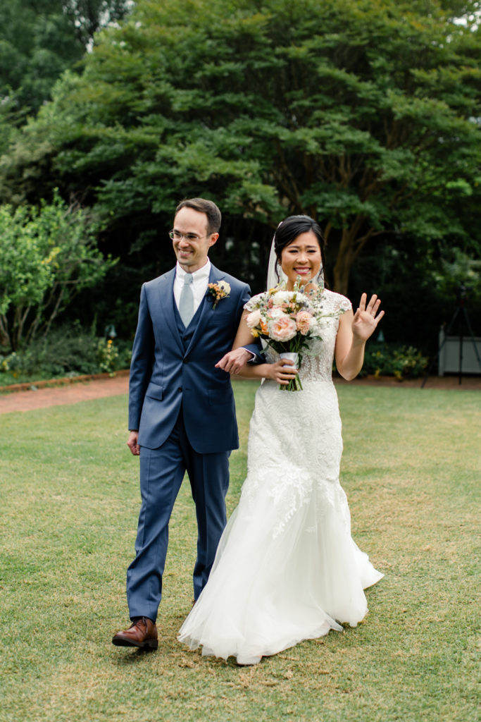 Bride in white lace dress and pink and white bouquet walking down aisle holding hands with groom in blue suit at Daniel Stowe Botanical wedding venue. Photographed by Charlotte wedding photography.