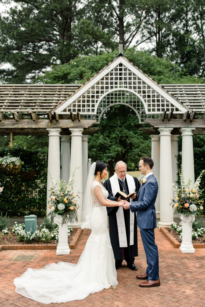 Bride in white lace gown holding hands with groom in blue suit under white alter at the Daniel Stowe Garden wedding venue.