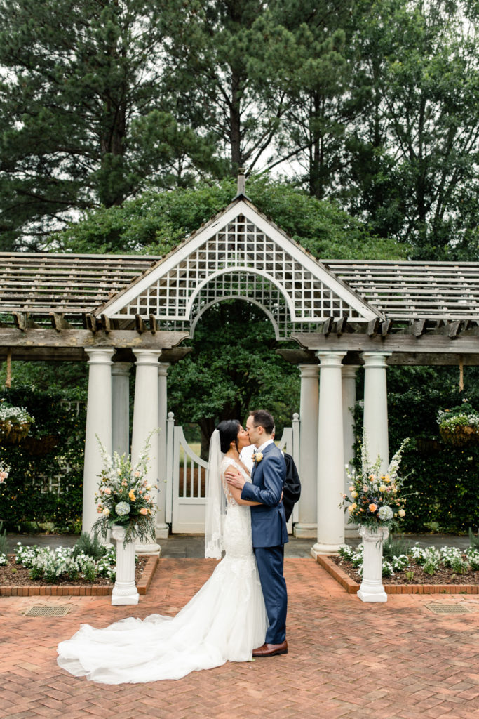 Bride in white lace gown kissing with groom in blue suit under white alter at the Daniel Stowe Garden wedding venue.