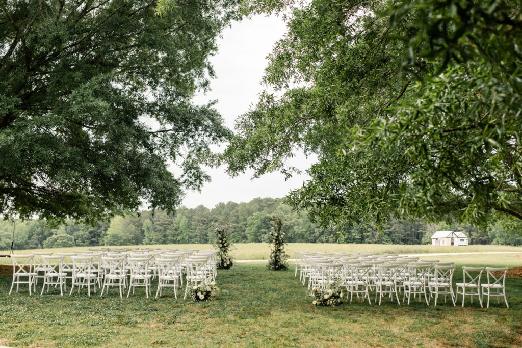 Wedding ceremony set up with one hundred white chairs with greenery and white flower arbor at The Meadows Raleigh wedding venue in Raleigh NC. Photographed by Charlotte wedding photographer.