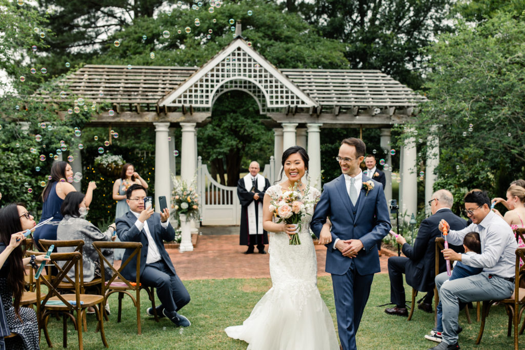 Bride in white lace dress and pink and white bouquet walking down aisle smiling with groom in blue suit at Daniel Stowe Botanical wedding venue. Photographed by Charlotte wedding photography.