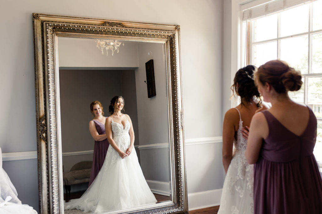 Bride in white lace dress looking at herself in gold mirror smiling with her bridesmaid in purple dress at SePark Mansion Gastonia Wedding Venue. Photographed by Charlotte wedding photographer.