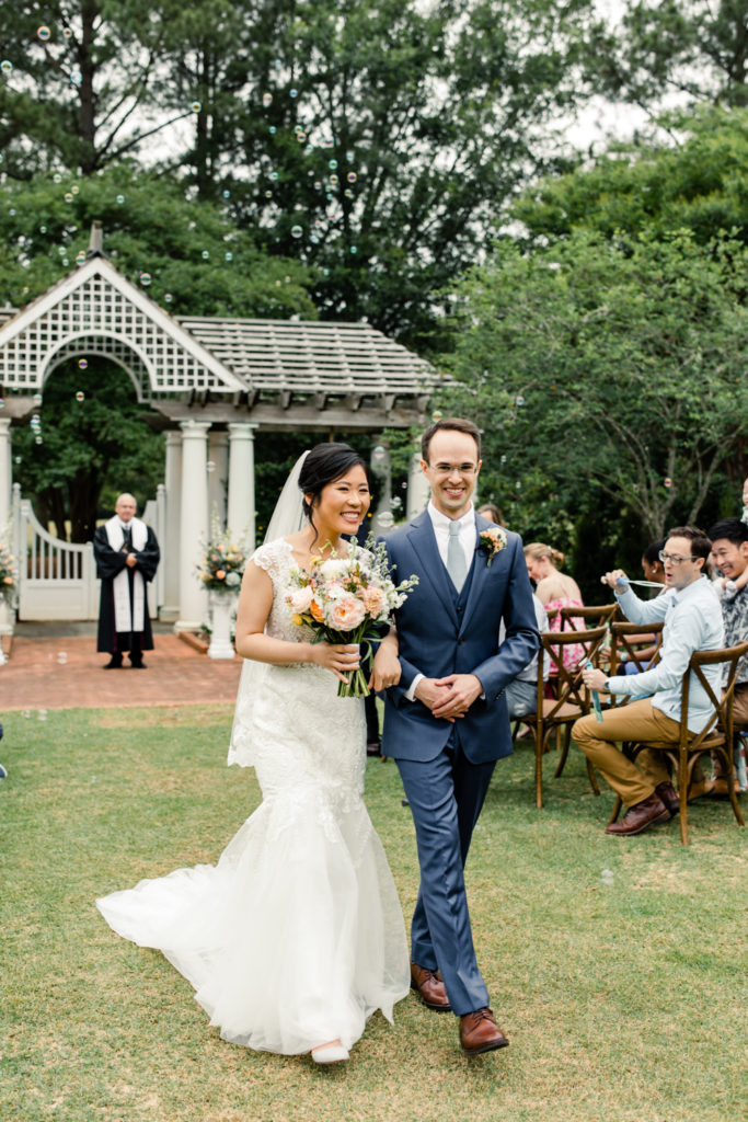 Bride in white lace dress and pink and white bouquet walking down aisle smiling with groom in blue suit at Daniel Stowe Botanical wedding venue. Photographed by Charlotte wedding photography.