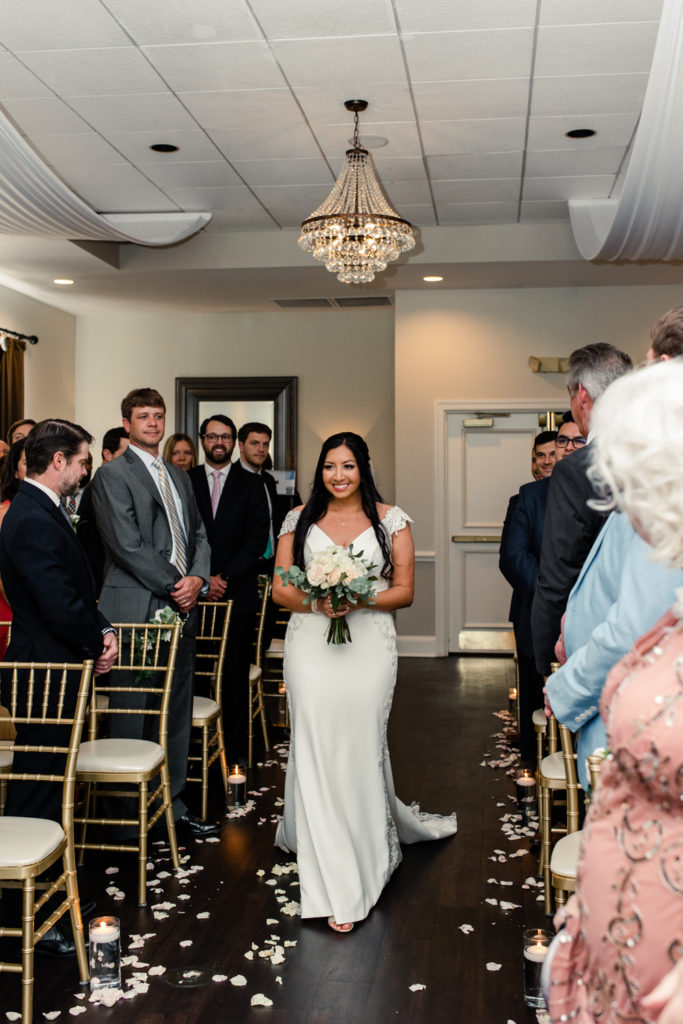 Bride with brown long hair in a white wedding dress holding a white bouquet walking down ceremony aisle at Northstone Country Club in Charlotte. Photographed by Charlotte wedding photographer.