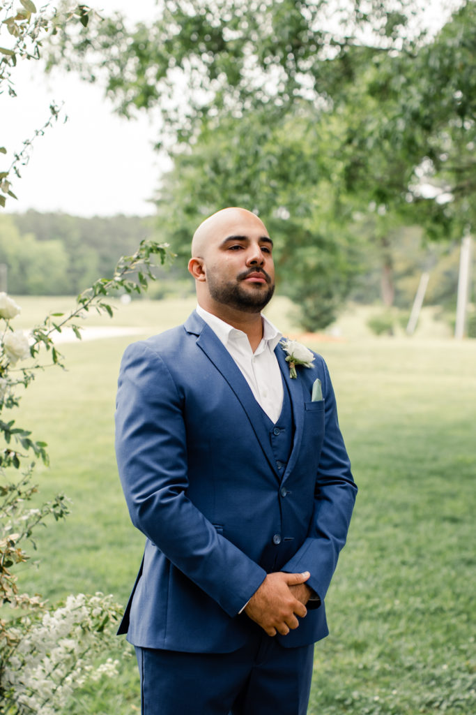 Groom, in blue suit and white boutonnière, watching his bride walk down the aisle at The Meadows Raleigh wedding venue in Raleigh NC. Photographed by Charlotte wedding photographer.