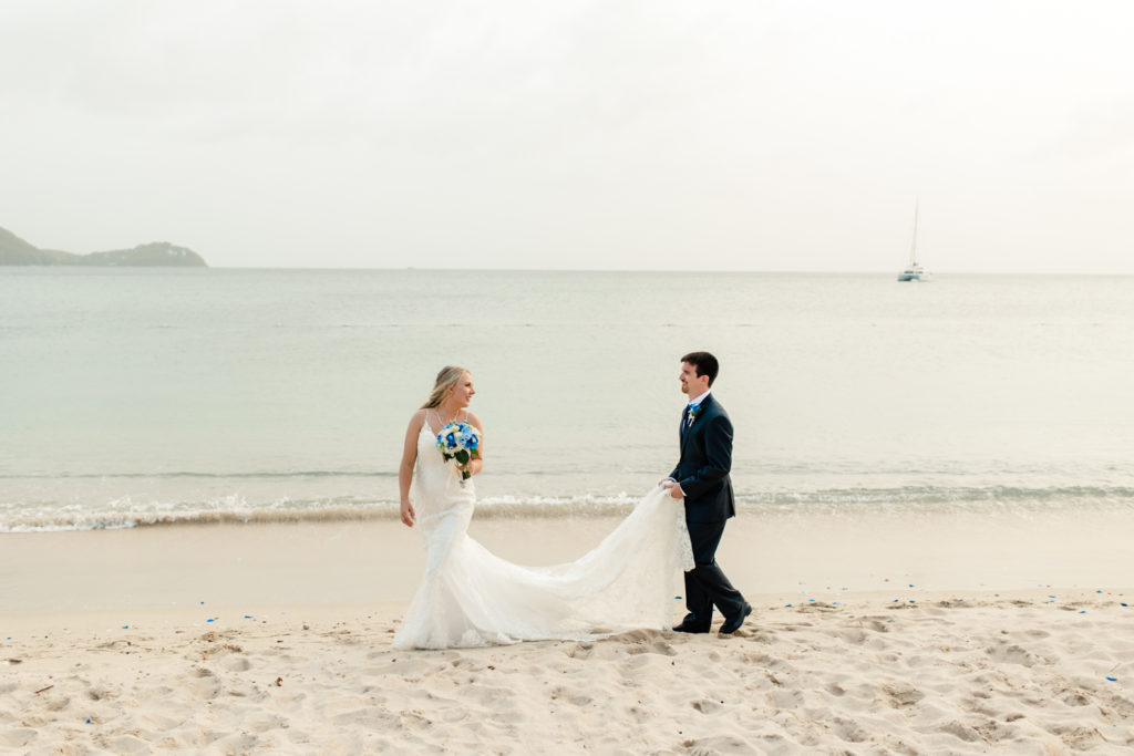 Bride walking on the coast of St. Lucia while the groom holds the bride's dress train at the St. Lucia Grande Sandals Resort. Photographed by Charlotte wedding photographer.