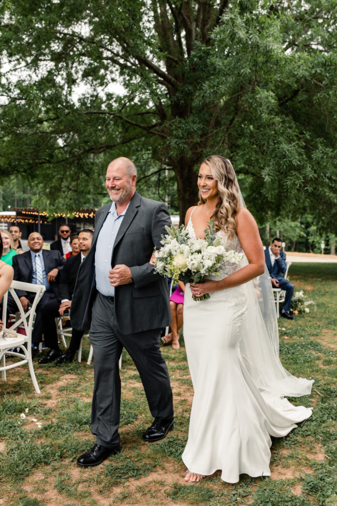 Bride, in white gown and white bouquet, walking down aisle with father at The Meadows Raleigh wedding venue in Raleigh NC. Photographed by Charlotte wedding photographer.