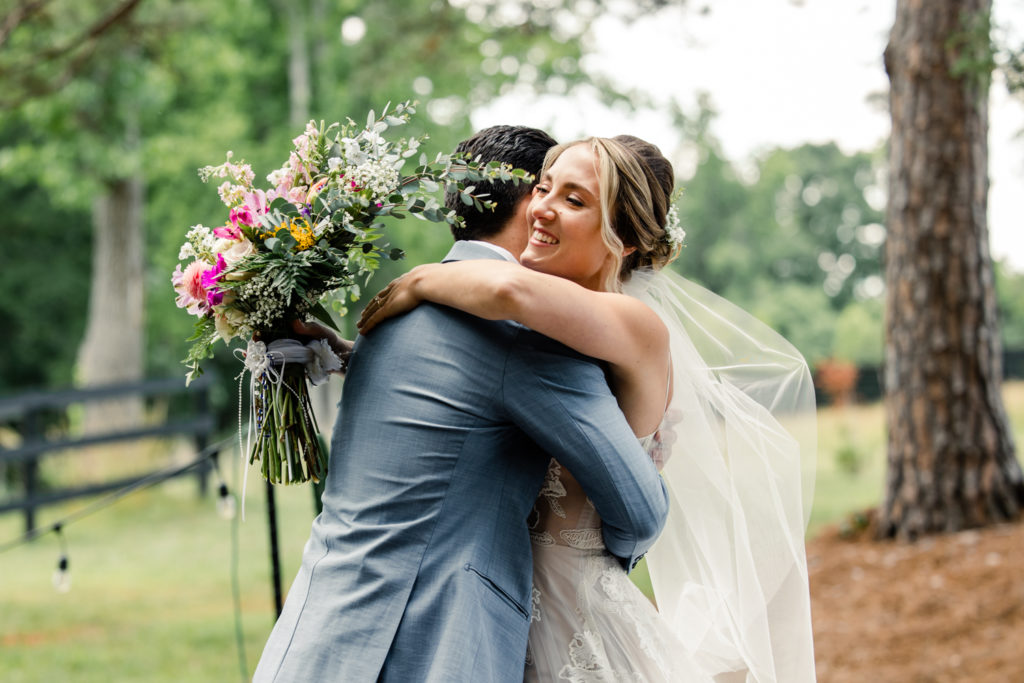 Bride in white lace dress holding green floral bouquet embraces groom in grey suit at Bansen Farms wedding venue in Charlotte NC.