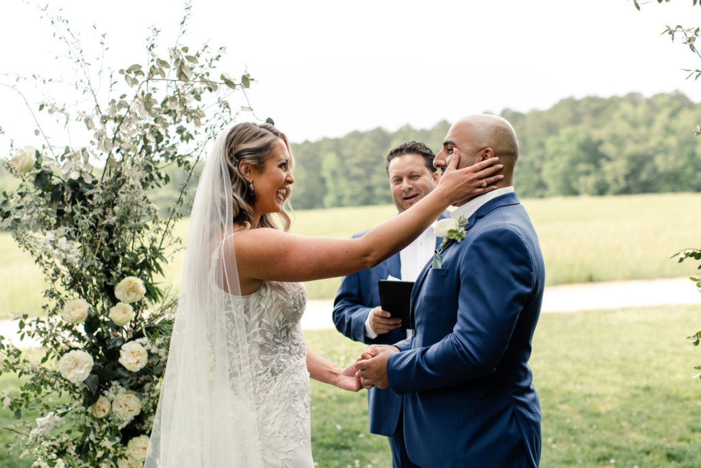 Bride, in white dress and veil, wiping the tear off her groom's face during ceremony at The Meadows Raleigh wedding venue in Raleigh NC. Photographed by Charlotte wedding photographer.