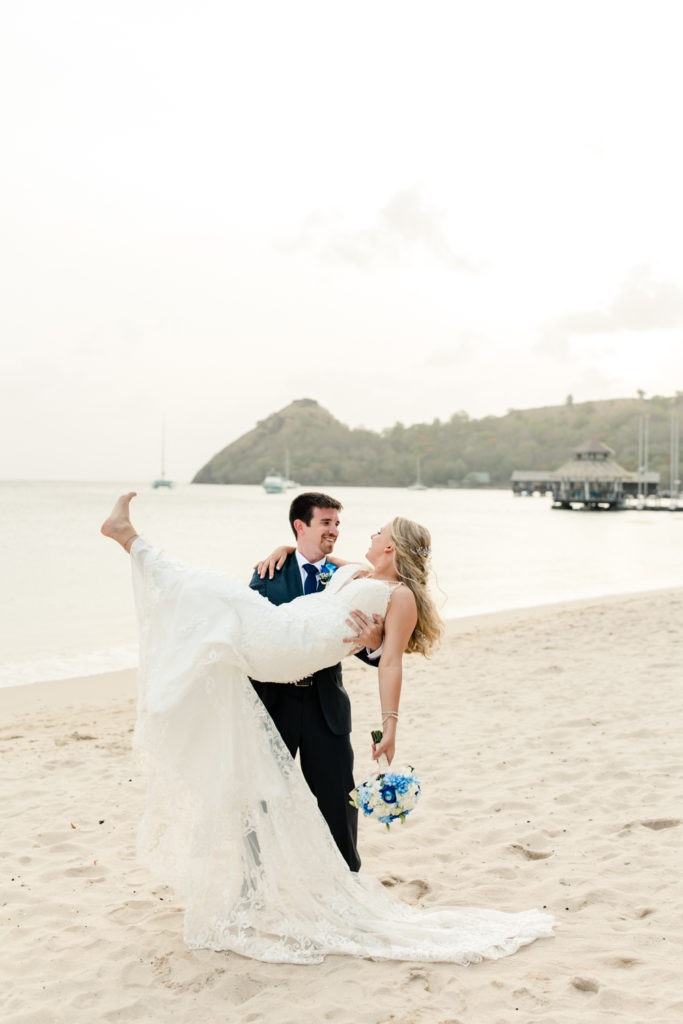 Groom, in black suit, picking up his bride, in white dress, at the St. Lucia Grande Sandals Resort. Photographed by Charlotte wedding photographer.