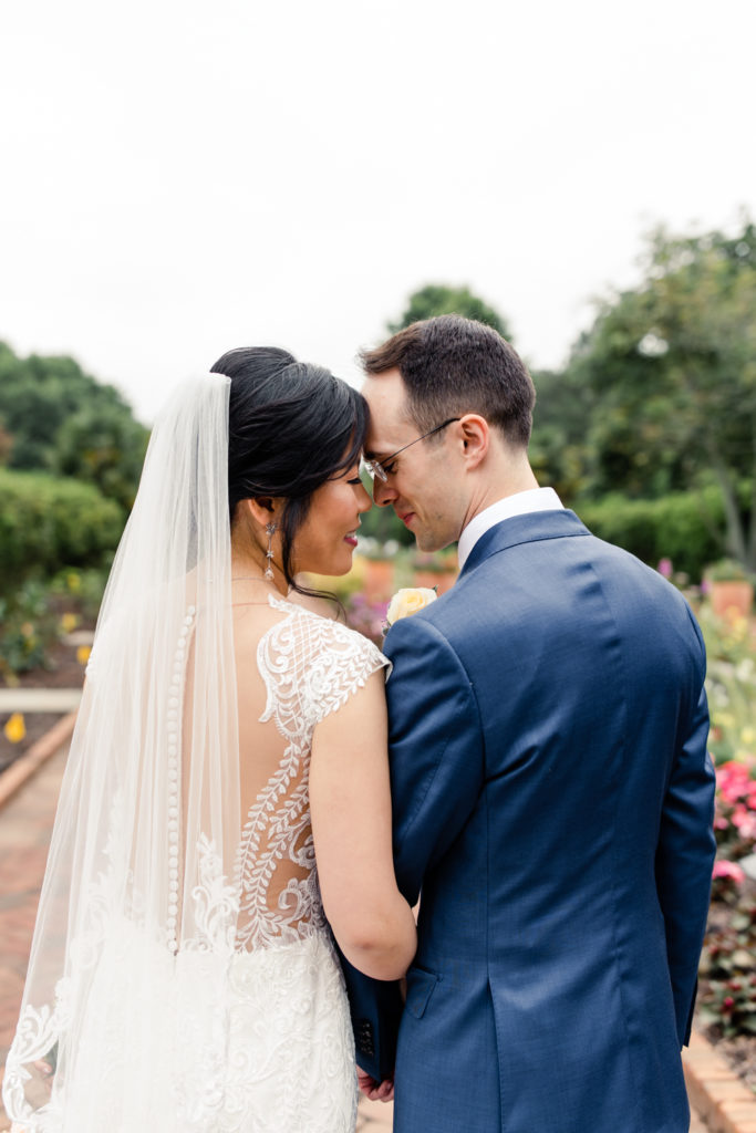 Bride in white lace wedding dress going nose to nose with her groom in a blue suit at Daniel Stowe Botanical Garden Wedding Venue. Photographed by Charlotte wedding photography.