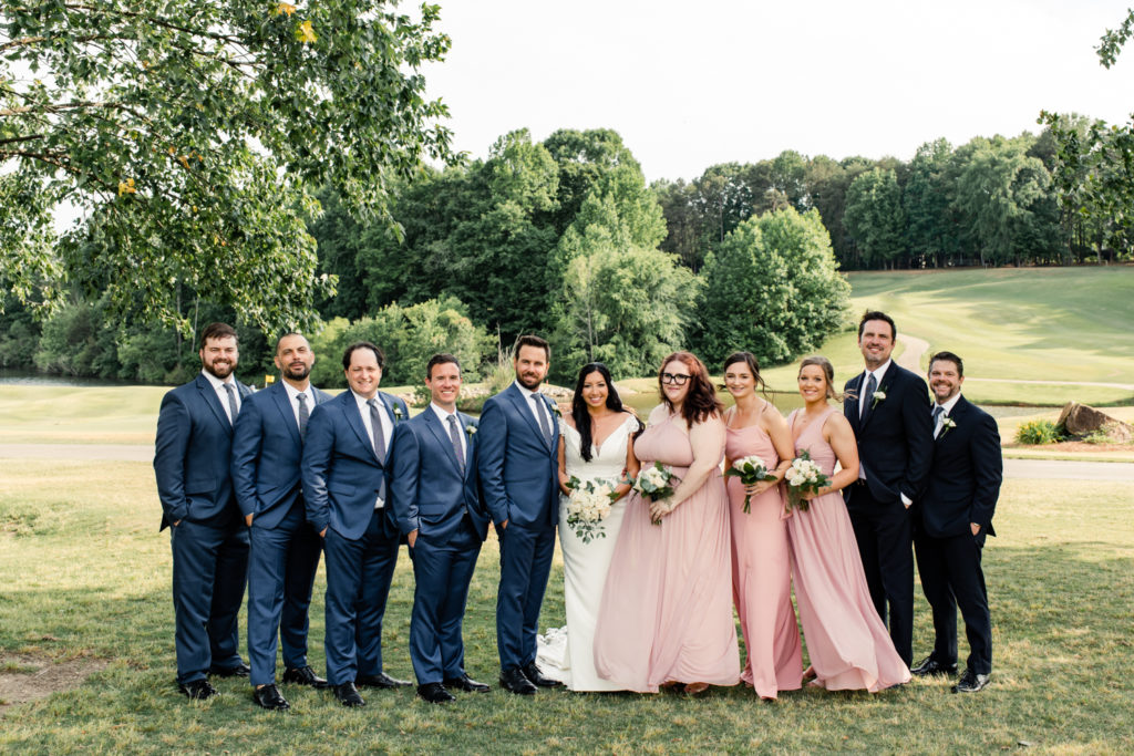 Bride in white dress and groom in blue suit stand with wedding party on golf course