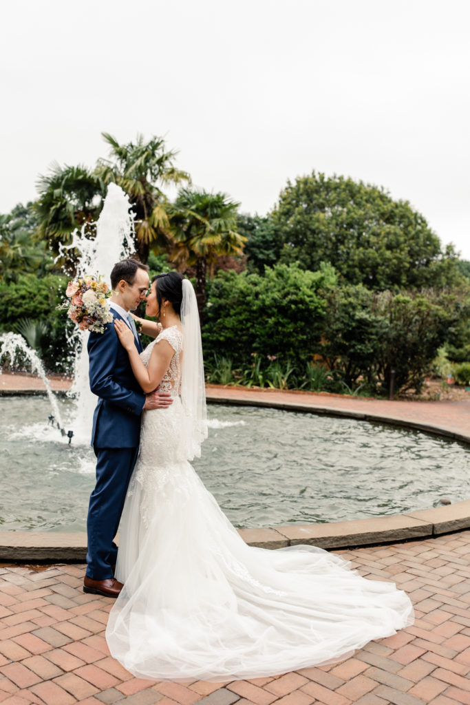 Bride in white lace wedding dress going nose to nose with her groom in a blue suit stand in front of a fountain at Daniel Stowe Botanical Garden Wedding Venue. Photographed by Charlotte wedding photography.