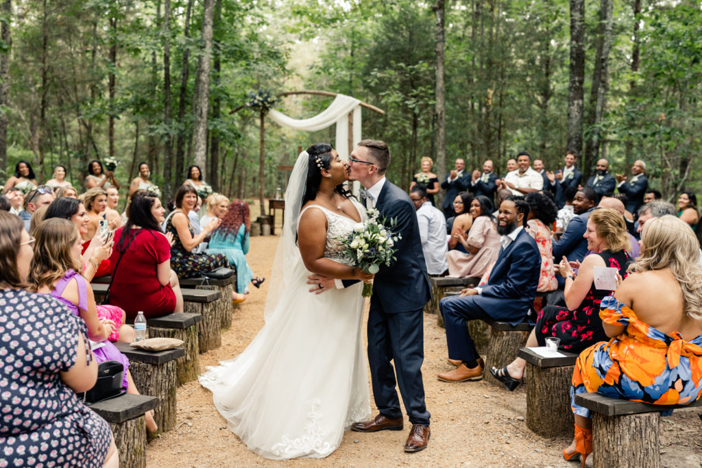 Bride, in white gown, kissing groom, in navy suit, in the middle of ceremony aisle at Carolina Country weddings venue in Charlotte.