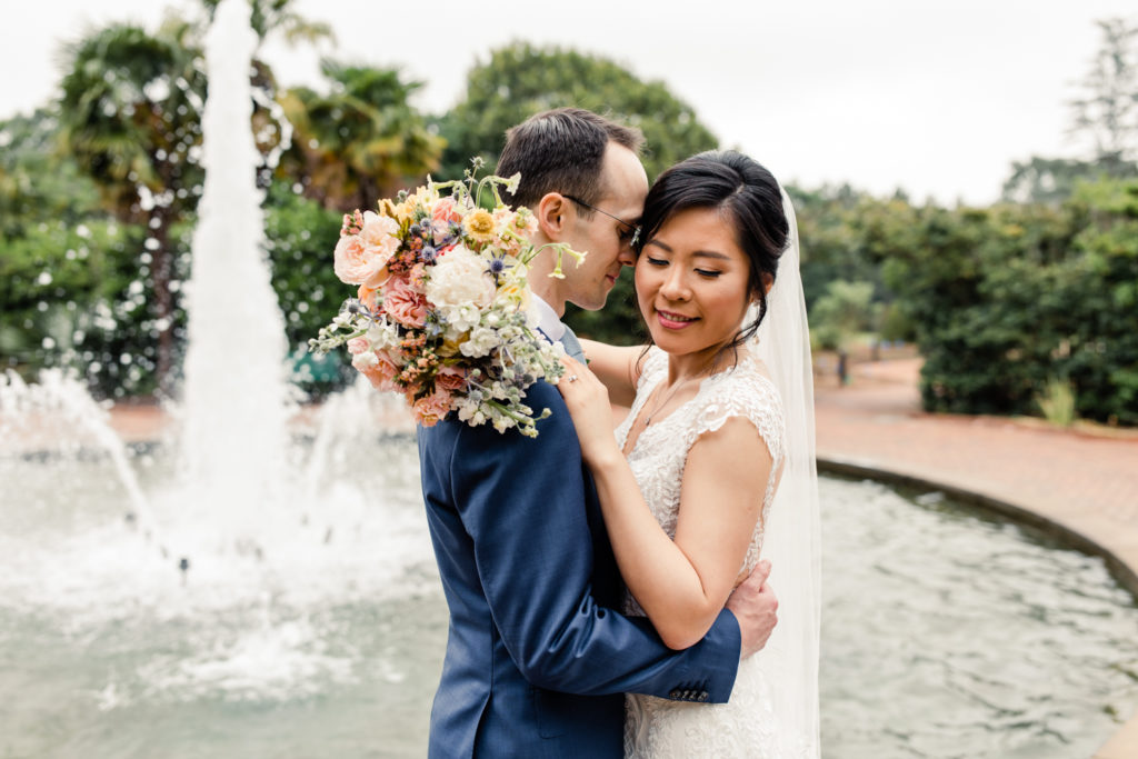 Bride in white lace gown and veil hugging groom with brown hair in blue suit at Daniel Stowe Botanical Gardens in Charlotte photographed by Stephanie Bailey Photography.