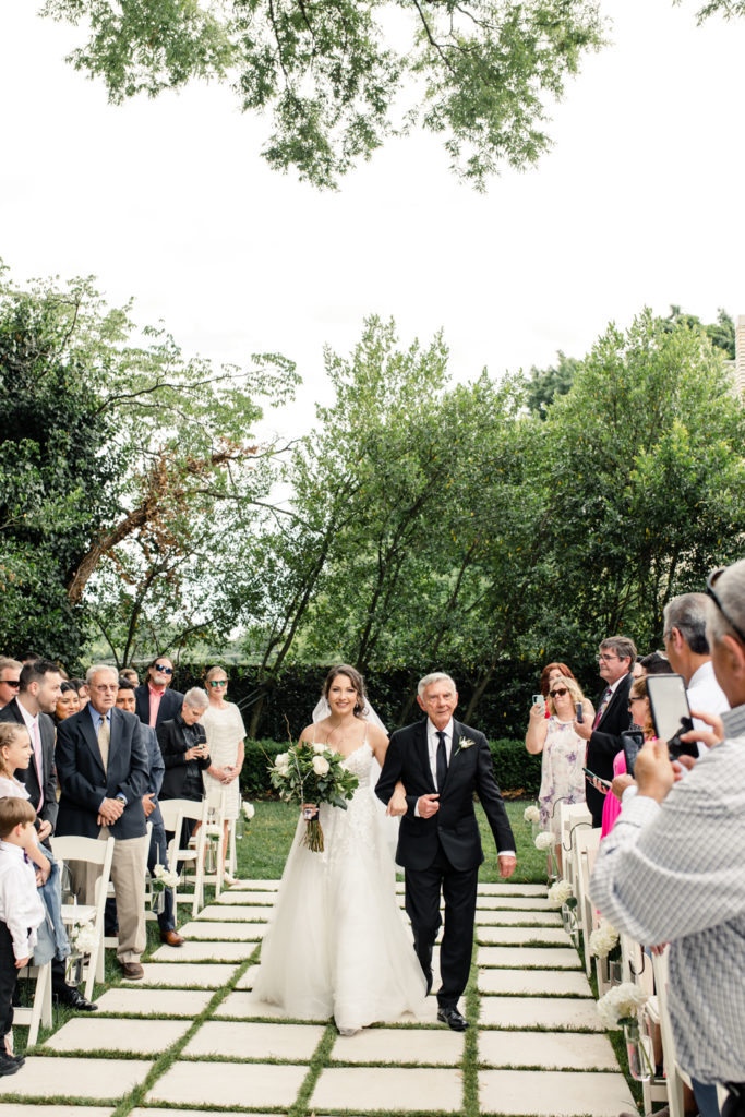 Bride wearing white lace gown holding white and green flowers walks down aisle with dad in black suit and tie at SePark Mansion Gastonia Wedding Venue. Photographed by Charlotte wedding photographer.