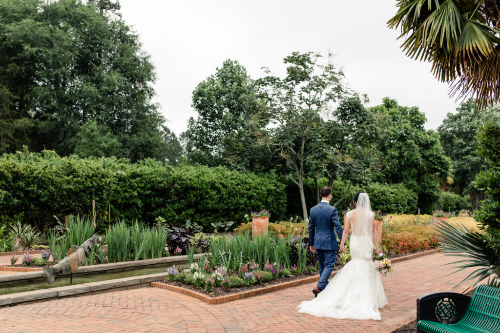 Bride in white lace dress and pink and white bouquet walking holding hands with groom in blue suit at Daniel Stowe Botanical wedding venue. Photographed by Charlotte wedding photography.