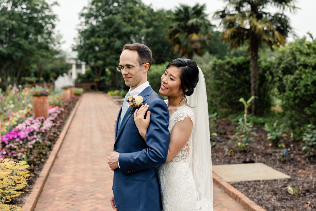 Bride in white lace gown and veil hugging groom with brown hair in blue suit at Daniel Stowe Botanical Gardens in Charlotte photographed by Stephanie Bailey Photography.