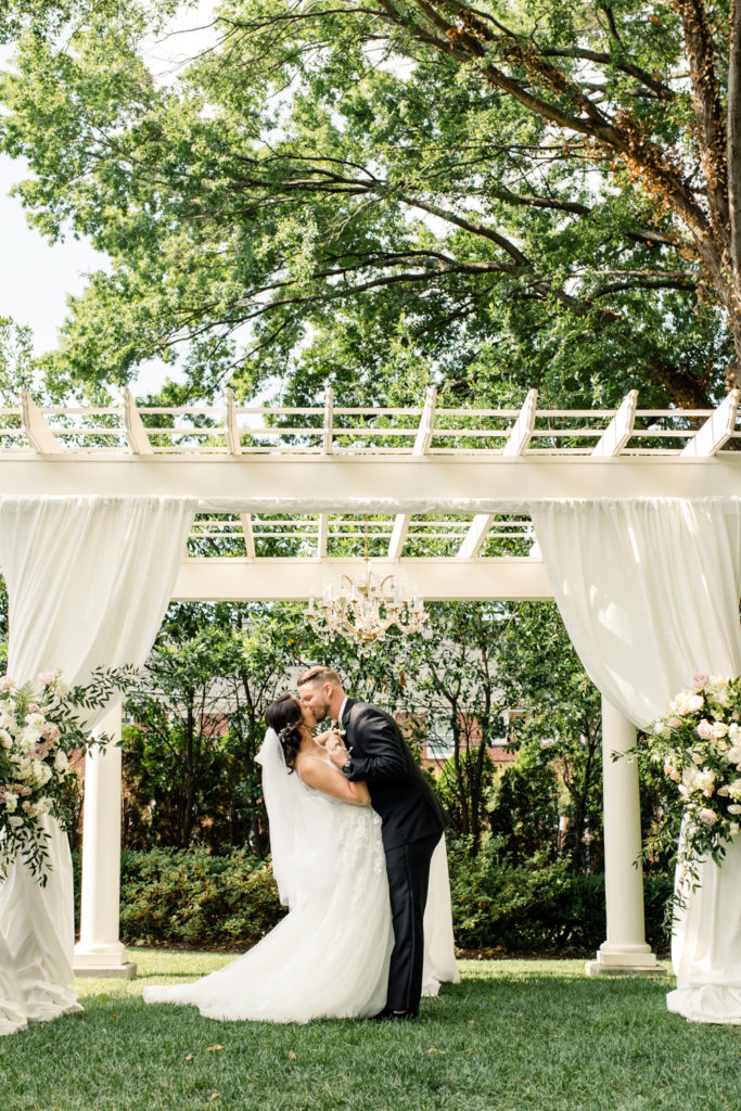 Groom in black suit kissing his bride in a white gown and veil under a white arbor at SePark Mansion Gastonia Wedding Venue. Photographed by Charlotte wedding photographer.