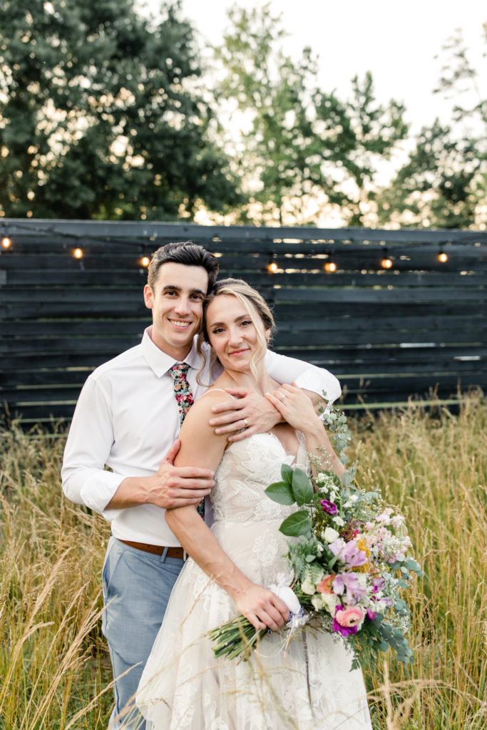Groom in white button down and floral tie hugging bride in white dress and white, pink and green bouquet at Bansen Farms wedding venue in Charlotte NC.