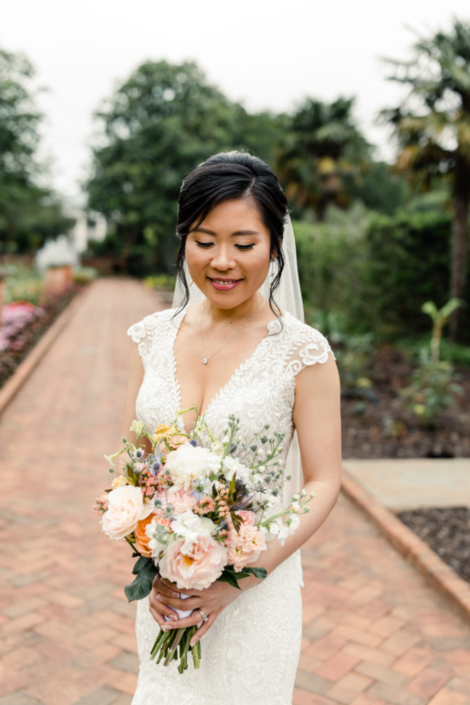 Bride in white lace dress holding pink and white bouquet smiling at flowers at the Daniel Stowe Botanical Garden wedding venue in Charlotte.