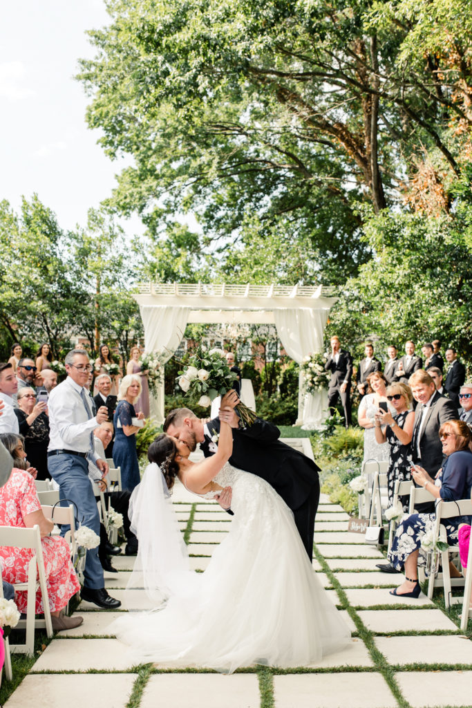 Groom in black suit kissing his bride in a white gown and veil in the middle of the ceremony aisle at SePark Mansion Gastonia Wedding Venue. Photographed by Charlotte wedding photographer.