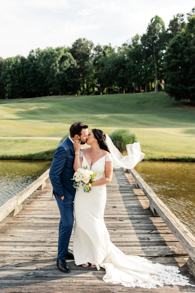 Bride in white wedding dress and veil kissing groom in blue suit on golf course bridge at Northstone Country Club in Charlotte. Photographed by Charlotte wedding photographer.