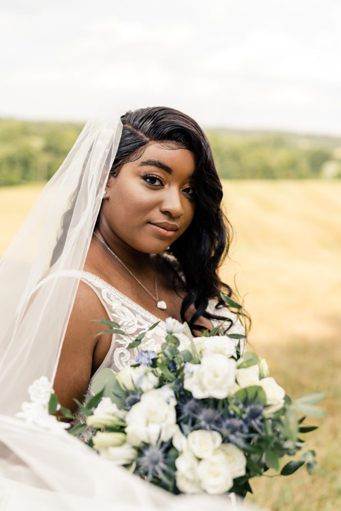 Bride, in white lace dress and veil, holding white and green bouquet in a field at Carolina Country weddings venue in Charlotte.