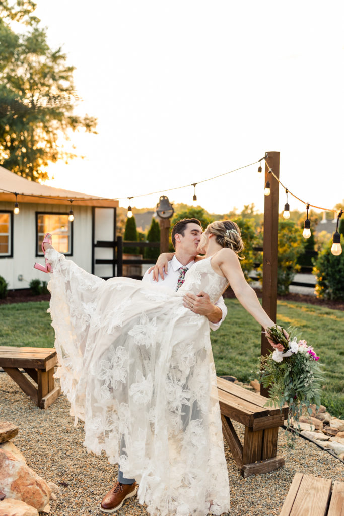 Groom in white shirt and grey pants carrying and kissing bride in white lace dress during sunset at Bansen Farms wedding venue in Charlotte NC.