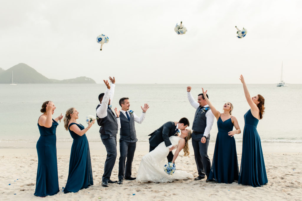 Bride, in white dress, kissing groom, in black suit, on the beach while wedding party throws flowers at the St. Lucia Grande Sandals Resort. Photographed by Charlotte wedding photographer.