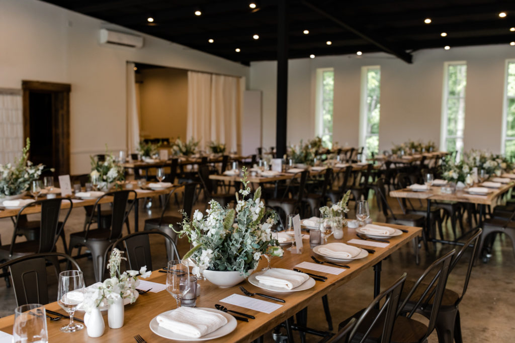 Wedding reception details of wood rustic tables with white and green bouquet table scapes at The Meadows Raleigh wedding venue in Raleigh NC. Photographed by Charlotte wedding photographer.