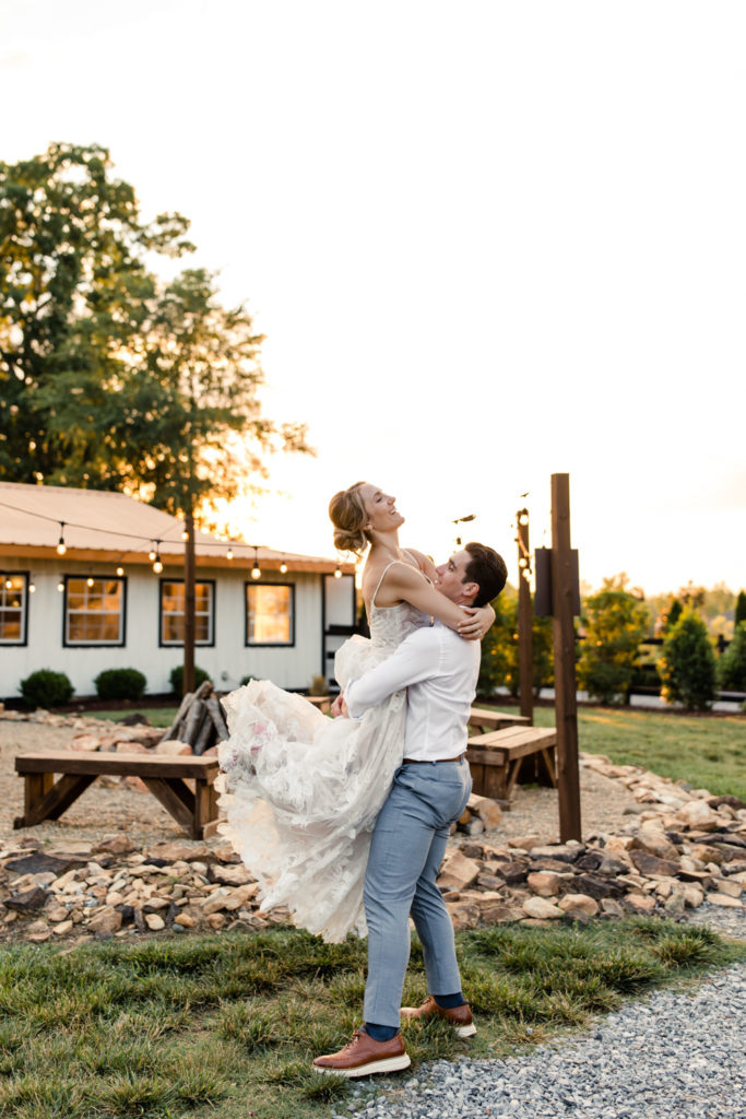 Groom in white shirt and grey pants carrying bride in white lace dress during sunset at Bansen Farms wedding venue in Charlotte NC.