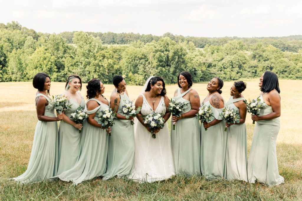 Bride, in white dress, laughing with eight bridesmaids, in green dresses at Carolina Country weddings venue in Charlotte.