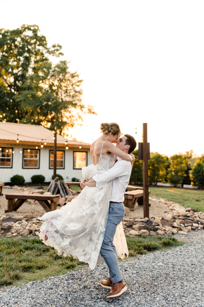 Groom in white shirt and grey pants carrying bride in white lace dress during sunset at Bansen Farms wedding venue in Charlotte NC.