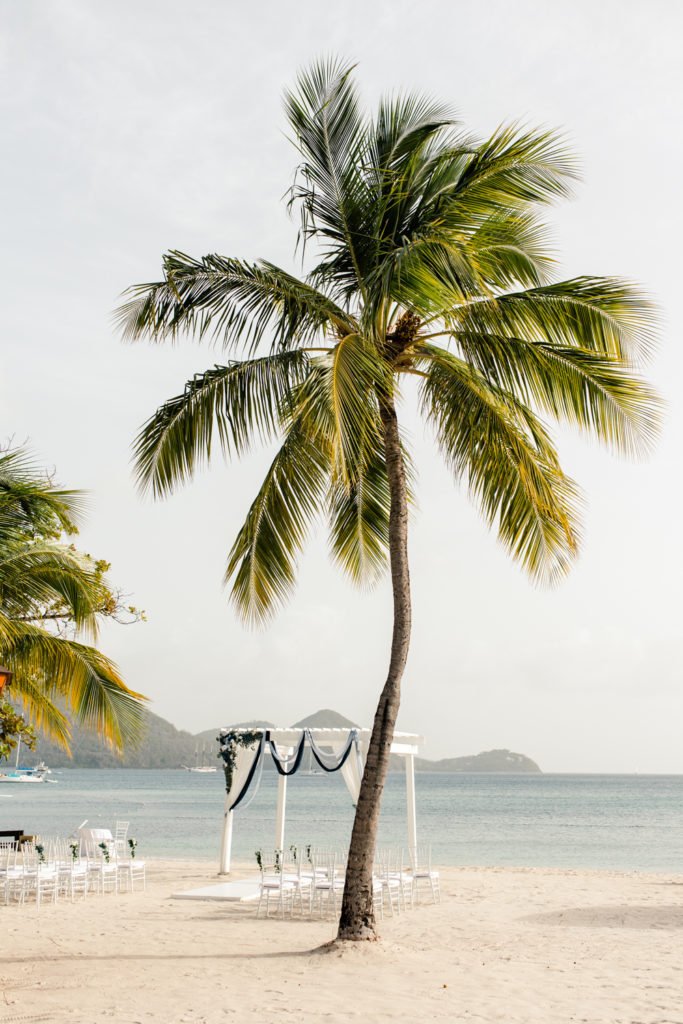 Ceremony set up on the beach of St Lucia with thirty white chairs and arbor surrounded by large palm chairs.